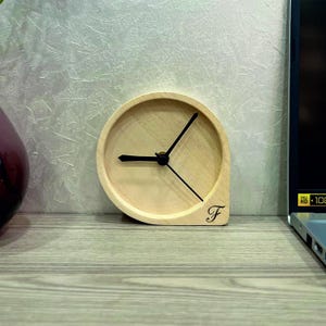 May include: A wooden clock with black hands and a simple design. The clock is shaped like a teardrop and has the letter "F" engraved on the side.