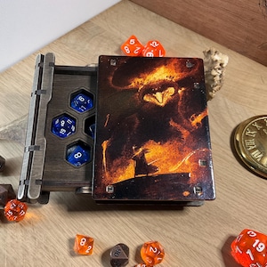 Puede incluir: Una caja de madera con una tapa con bisagras, que contiene un juego de dados azules. La tapa presenta una imagen impresa de una escena de fuego con una figura oscura en el centro. La caja está rodeada de dados naranjas.