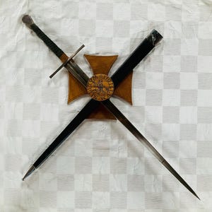 May include: A medieval sword and scabbard are crossed over a wooden emblem with a circular design. The sword has a silver blade and a dark green wrapped handle. The scabbard is black. The emblem is brown with a gold-coloured circular centre.