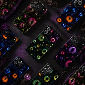 May include: A collection of rectangular smartphone cases featuring a repeating pattern of various reptilian eyes in vibrant colors including green, purple, blue, and orange. Each case has a camera cutout and a glossy finish with dark shadows.