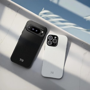 May include: Two smartphone cases, one black and one white, with a textured, striped design. The black case has the initials "YN" and the white case has "HI". Both cases have camera cutouts and are angled on a light surface.