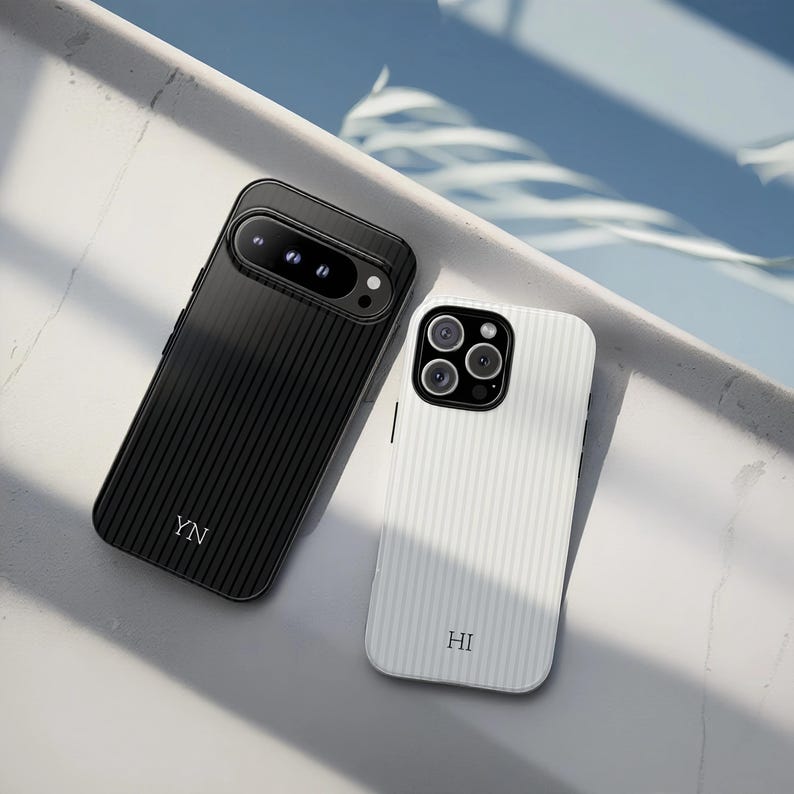 May include: Two smartphone cases, one black and one white, with a textured, striped design. The black case has the initials "YN" and the white case has "HI". Both cases have camera cutouts and are angled on a light surface.