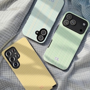 May include: Three phone cases with vertical striped patterns in pastel colors: light blue, mint green, and yellow. Each case has a different set of initials: "KM", "OL", and "YE". The cases are lying on a wrinkled, light gray fabric.