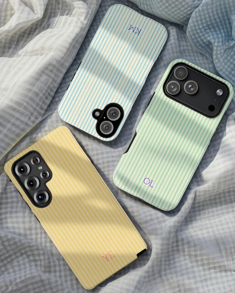 May include: Three phone cases with vertical striped patterns in pastel colors: light blue, mint green, and yellow. Each case has a different set of initials: "KM", "OL", and "YE". The cases are lying on a wrinkled, light gray fabric.