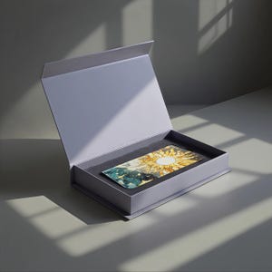 Op de afbeelding: Een rechthoekige, lichtpaarse doos met een magnetische sluiting, open om een kaart met een zon- en hemels ontwerp te onthullen. De kaart is genesteld in zwart schuim. De doos staat op een grijs oppervlak.