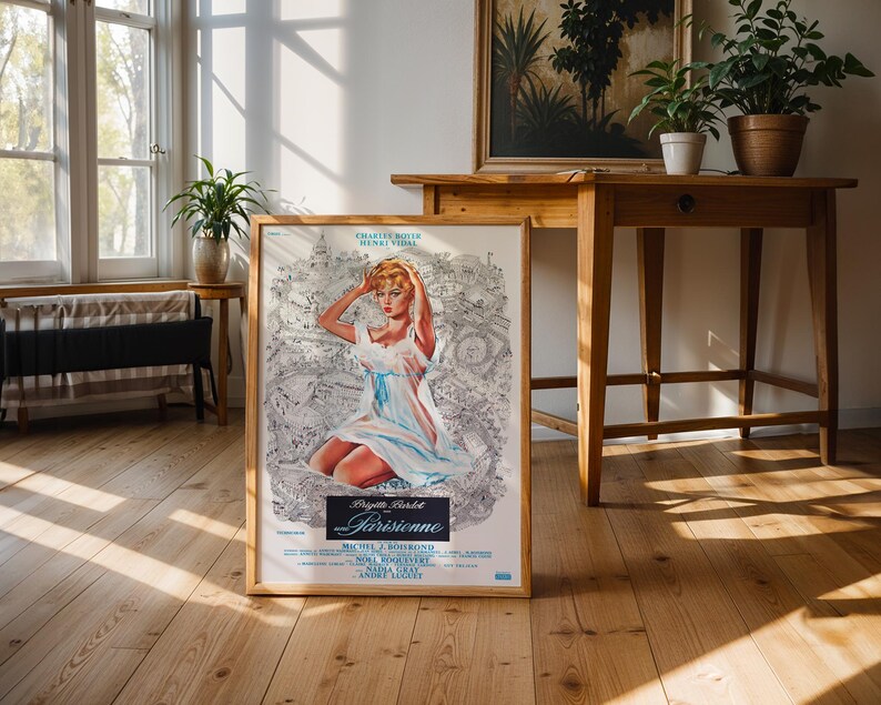 May include: A framed vintage movie poster featuring Brigitte Bardot in a light blue dress. The poster has a light wood frame and is displayed on a wooden floor. The poster's title is "Parisienne".