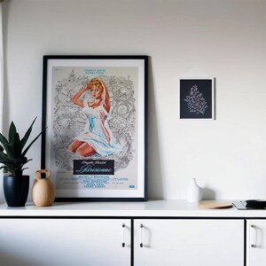 May include: A framed vintage movie poster featuring a woman in a light blue dress, with the title "Parisienne." A small black and white art print hangs nearby. A black potted plant and decorative vases sit on a white cabinet.