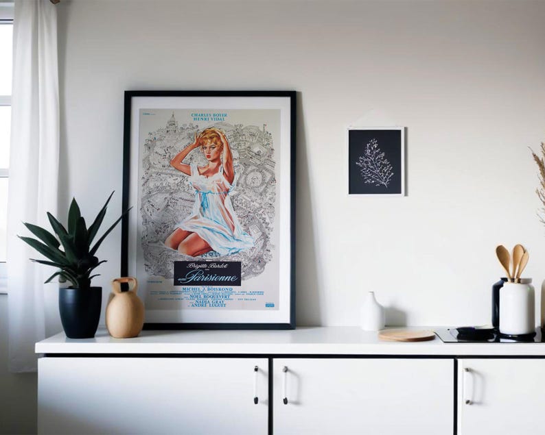 May include: A framed vintage movie poster featuring a woman in a light blue dress, with the title "Parisienne." A small black and white art print hangs nearby. A black potted plant and decorative vases sit on a white cabinet.