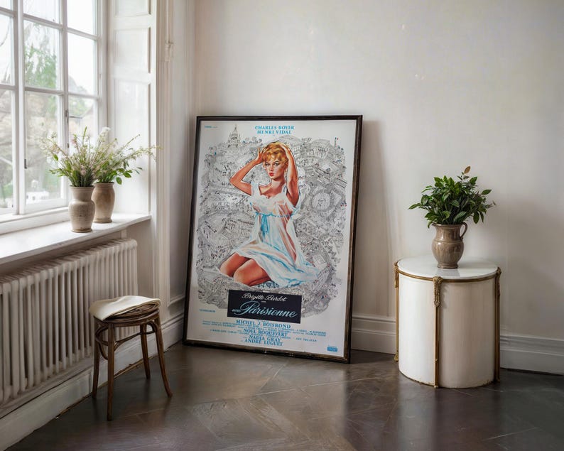 May include: A framed vintage movie poster featuring a woman in a white dress, with the title "Parisienne". The poster is leaning against a white wall. A small, round side table with a marble top and a vase of greenery are next to the poster.