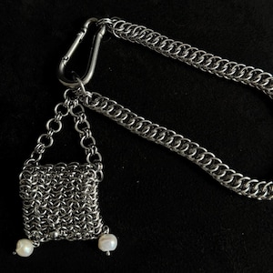 Könnte beinhalten: Ein silberfarbenes Kettenhemd-Accessoire mit einem quadratischen Beutel aus ineinandergreifenden Metallringen, der an einer langen Kette an einem Karabinerhaken hängt. Zwei kleine, weiße, unregelmäßig geformte Perlen sind an den unteren Ecken befestigt.