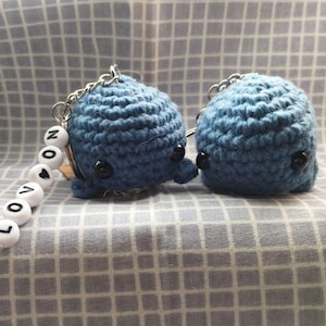 May include: Two small blue crocheted whale keychains with black eyes. One keychain has a white beaded charm that spells "LOVE YOU".