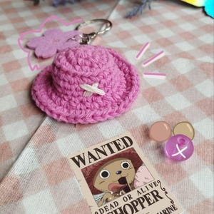 Puede incluir: Llavero de sombrero de crochet rosa con un diseño de cruz blanca. El llavero está unido a un anillo de llave plateado. El sombrero está sobre una tela a cuadros rosa y blanca. Un cartel de búsqueda de Chopper del anime One Piece está en el fondo.