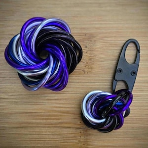 May include: Two metal keychains with a knot design. One keychain is a cluster of interlocking rings in shades of purple, silver, and black. The other keychain has a similar knot design attached to a black carabiner clip.