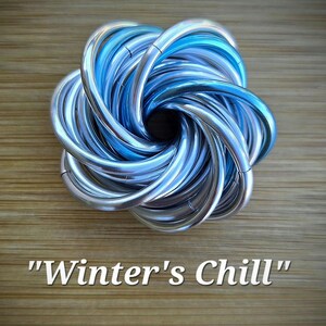 May include: A metallic sculpture with a swirling, interwoven design in shades of silver and blue. The sculpture is set against a wooden background and features the text "Winter's Chill".