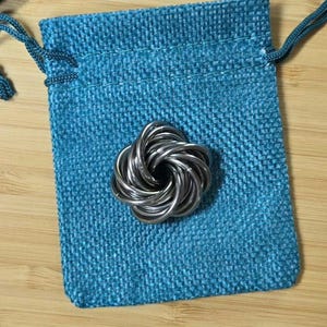May include: A silver-colored metal fidget toy in a knot design rests on a teal blue drawstring bag. The bag is made of a textured fabric and sits on a light brown wooden surface. The fidget toy is made of intertwined metal loops.