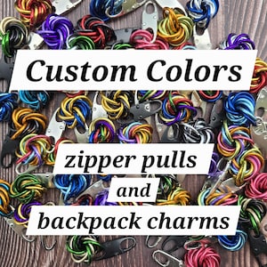 May include: An assortment of colorful zipper pulls and backpack charms in various hues, including green, purple, red, and blue. The image features the text "Custom Colors" and "zipper pulls and backpack charms" in a white rectangular box.