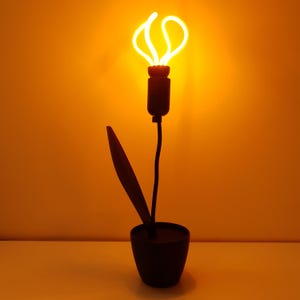 Może przedstawiać: Czarna metalowa lampa stołowa w kształcie kwiatu z jednym liściem i żarówką w środku. Żarówka jest włączona i świeci na żółto.