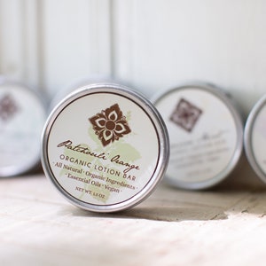 May include: A silver tin with a white label that reads "Patchouli Orange Organic Lotion Bar" with the text "All Natural Organic Ingredients Essential Oils Vegan Net Wt. 1.4 oz".