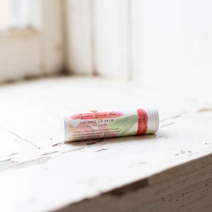 May include: A white tube of organic lip balm with a pink and green watercolor design. The tube is labeled "Lyla Green Tea Organic Lip Balm Net Wt .15 oz (4.25g)"