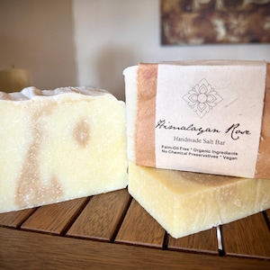 Op de afbeelding: Twee repen handgemaakte Himalayazoutzeep van Himalayan Rove. De zeep is lichtgeel met bruine swirls. De bovenste reep heeft een bruin papieren etiket met de tekst "Himalayan Rove Handgemaakte Zoutzeep Palmolievrij * Biologische Ingredi&euml;nten Geen Chemische Conserveermiddelen * Vegan".