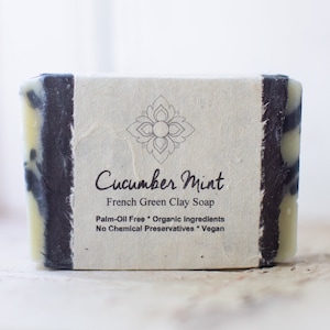 May include: A bar of Cucumber Mint French Green Clay Soap. The soap is palm-oil free, made with organic ingredients, and is vegan. It is packaged in a white paper wrapper with black edges.