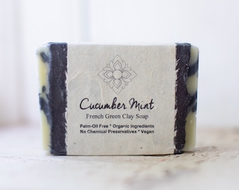 May include: A bar of Cucumber Mint French Green Clay Soap. The soap is palm-oil free, made with organic ingredients, and is vegan. It is packaged in a white paper wrapper with black edges.