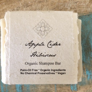 May include: A white bar of shampoo with the text "Apple Cider Hibiscus Organic Shampoo Bar Palm-Oil Free * Organic Ingredients No Chemical Preservatives * Vegan" printed on it.