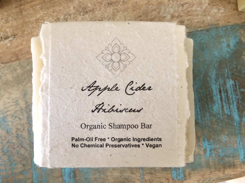 May include: A white bar of shampoo with the text "Apple Cider Hibiscus Organic Shampoo Bar Palm-Oil Free * Organic Ingredients No Chemical Preservatives * Vegan" printed on it.