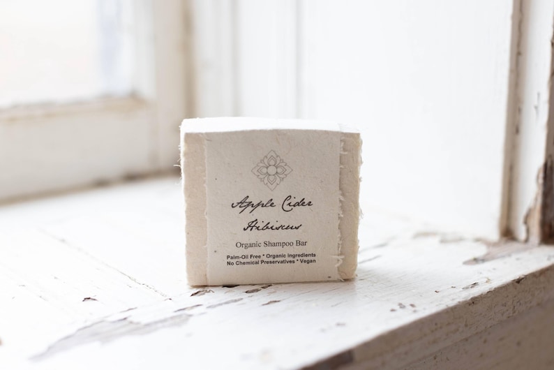 May include: A white bar of organic shampoo with the text "Apple Cider Hibiscus Organic Shampoo Bar" and "Palm-Oil Free * Organic Ingredients * No Chemical Preservatives * Vegan" printed on the label.