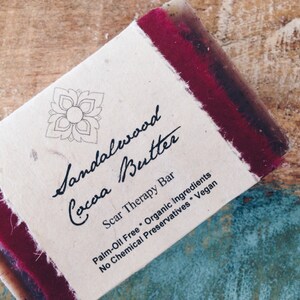 May include: A bar of sandalwood and cocoa butter soap with a red and white label that reads "Sandalwood Cocoa Butter Scar Therapy Bar Palm-Oil Free Organic Ingredients No Chemical Preservatives Vegan".