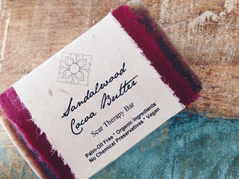 May include: A bar of sandalwood and cocoa butter soap with a red and white label that reads "Sandalwood Cocoa Butter Scar Therapy Bar Palm-Oil Free Organic Ingredients No Chemical Preservatives Vegan".
