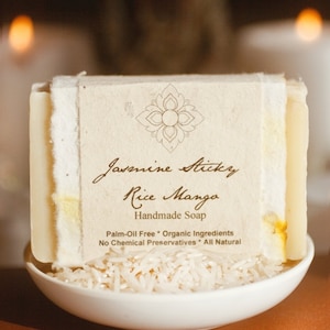 May include: A bar of handmade Jasmine Sticky Rice Manga soap, wrapped in white paper with a brown label. The label features a floral design and the text "Jasmine Sticky Rice Manga Handmade Soap Palm-Oil Free * Organic Ingredients No Chemical Preservatives * All Natural". The soap is sitting on a white bowl filled with white rice.