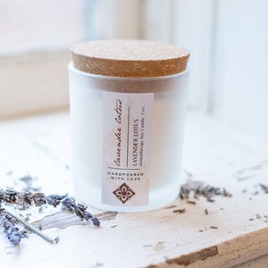 May include: A white frosted glass candle jar with a cork lid. The label reads "Lavender Lotus Aromatherapy Soy Candle, 7 oz.  Handpoured with Love."  The candle is sitting on a white wooden surface with dried lavender flowers.