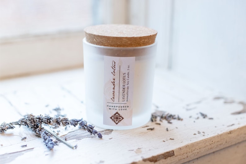 May include: A white frosted glass candle jar with a cork lid. The label reads "Lavender Lotus Aromatherapy Soy Candle, 7 oz.  Handpoured with Love."  The candle is sitting on a white wooden surface with dried lavender flowers.