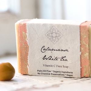 May include: A bar of white tea-scented soap with a pink and gold patterned wrapper. The wrapper has the text "Calamansi White Tea Vitamin C Face Soap Palm-Oil Free Organic Ingredients No Chemical Preservatives Vegan".
