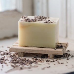 May include: A bar of white soap with dried lavender flowers on top, sitting on a wooden soap dish. The soap is on a white surface with dried lavender flowers scattered around it.
