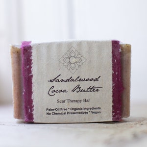 May include: A bar of sandalwood and cocoa butter soap with a brown and white label that reads "Sandalwood Cocoa Butter Scar Therapy Bar Palm-Oil Free Organic Ingredients No Chemical Preservatives Vegan".