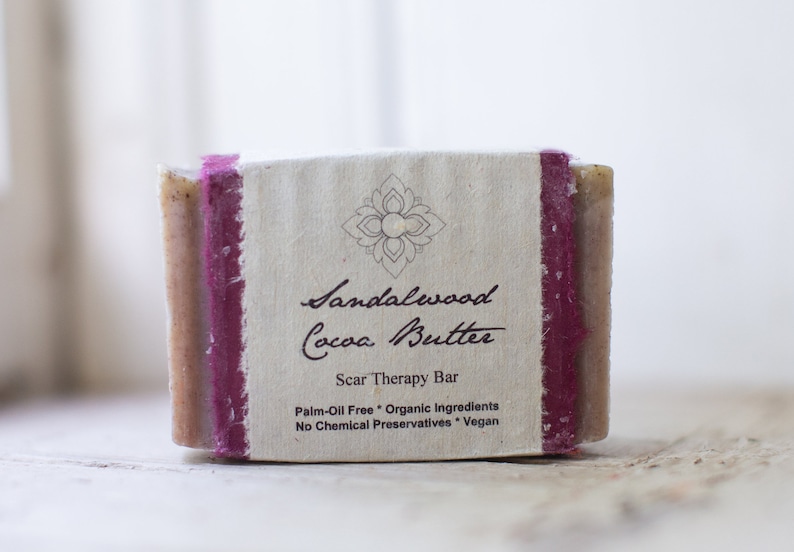 May include: A bar of sandalwood and cocoa butter soap with a brown and white label that reads "Sandalwood Cocoa Butter Scar Therapy Bar Palm-Oil Free Organic Ingredients No Chemical Preservatives Vegan".