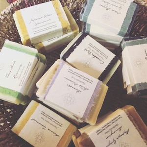 May include: A collection of handmade soap bars with various scents, including Japanese Citrine Chamomile, Cucumber Mint, Lotus Blossom, and Black Pepper. Each bar is wrapped in a decorative paper with a label listing the scent and ingredients. The soaps are displayed in a woven basket.