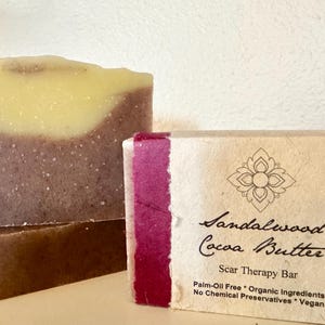 May include: Two bars of handmade soap, one with a swirl of light yellow and a base of purple, and the other with a label that reads "Sandalwood Cocoa Butter Scar Therapy Bar Palm-Oil Free * Organic Ingredients No Chemical Preservatives * Vegan"