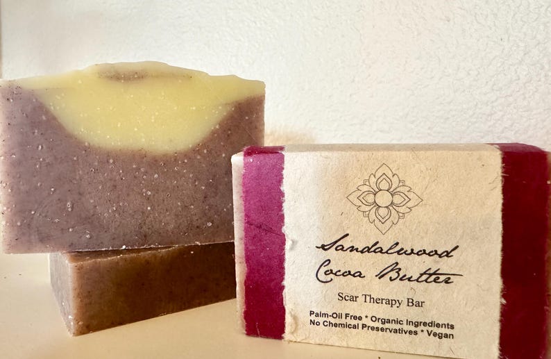 May include: Two bars of handmade soap, one with a swirl of light yellow and a base of purple, and the other with a label that reads "Sandalwood Cocoa Butter Scar Therapy Bar Palm-Oil Free * Organic Ingredients No Chemical Preservatives * Vegan"