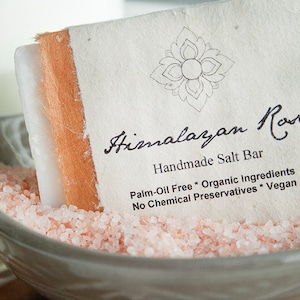 Op de afbeelding: Een witte handgemaakte Himalayazoutbar met de naam "Himalayan Rose Handmade Salt Bar Palm-Oil Free * Organic Ingredients No Chemical Preservatives * Vegan" in een kom met roze Himalayazout.
