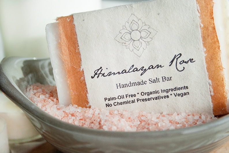 Op de afbeelding: Een witte handgemaakte Himalayazoutbar met de naam "Himalayan Rose Handmade Salt Bar Palm-Oil Free * Organic Ingredients No Chemical Preservatives * Vegan" in een kom met roze Himalayazout.