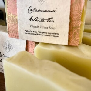 May include: Close-up of a stack of white soap bars with a pink and gold label that reads "Calamansi White Tea Vitamin C Face Soap Palm-Oil Free * Organic Ingredients No Chemical Preservatives * Vegan"