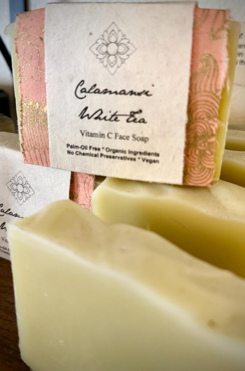 May include: Close-up of a stack of white soap bars with a pink and gold label that reads "Calamansi White Tea Vitamin C Face Soap Palm-Oil Free * Organic Ingredients No Chemical Preservatives * Vegan"