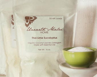 May include: A white resealable bag of Thai Lime Eucalyptus laundry detergent with a brown label that reads "Unearth Malee HOME" and "32-64 Loads". The bag is sitting on a white surface next to a small green bowl filled with white powder.