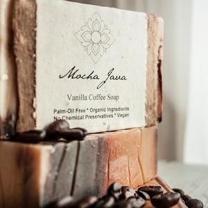 May include: Two bars of Mocha Java vanilla coffee soap, stacked on top of each other. The soap is brown and white with coffee beans scattered around the bars. The label reads "Mocha Java Vanilla Coffee Soap, Palm-Oil Free * Organic Ingredients, No Chemical Preservatives * Vegan."