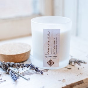 May include: A white frosted glass candle jar with a cork lid. The candle is labelled "Lavender Lotus" and "Aromatherapy Soy Candle, 7 oz." The label also says "Handpoured with Love". There are dried lavender flowers on a white wooden surface.