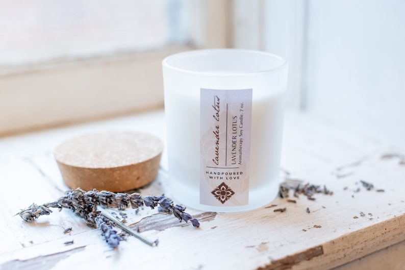 May include: A white frosted glass candle jar with a cork lid. The candle is labelled "Lavender Lotus" and "Aromatherapy Soy Candle, 7 oz." The label also says "Handpoured with Love". There are dried lavender flowers on a white wooden surface.