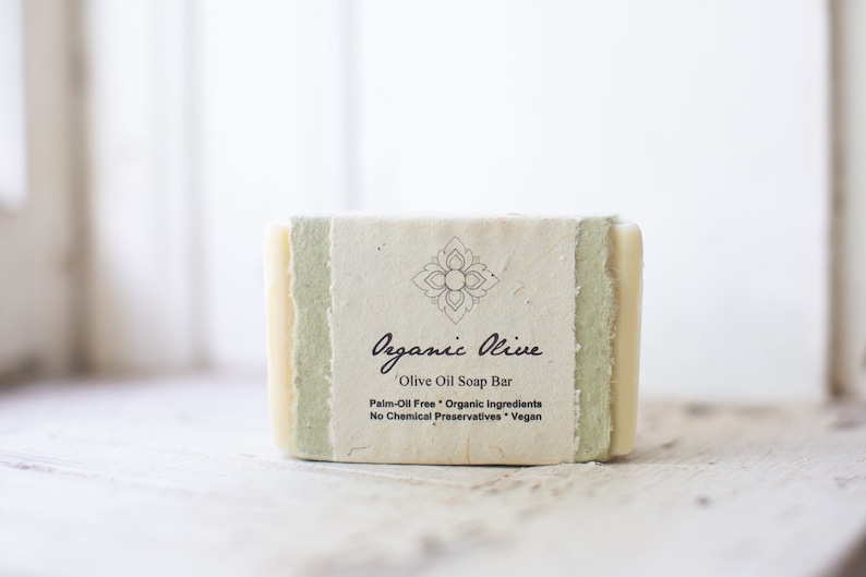 A single bar of cream-colored soap placed by a window. Soft morning light is shining on the soap. It's wrapped in two layers of flower-seeded paper. The first layer is sage green and the outer layer is off-white and has the label printed on it.